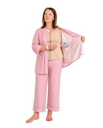 A woman wearing pink striped recovery  pajamas set with and a beige mastectomy tank top, holding open the striped top while demonstrating a clear plastic  tubing and surgical jp drain placement into the interior pocket.