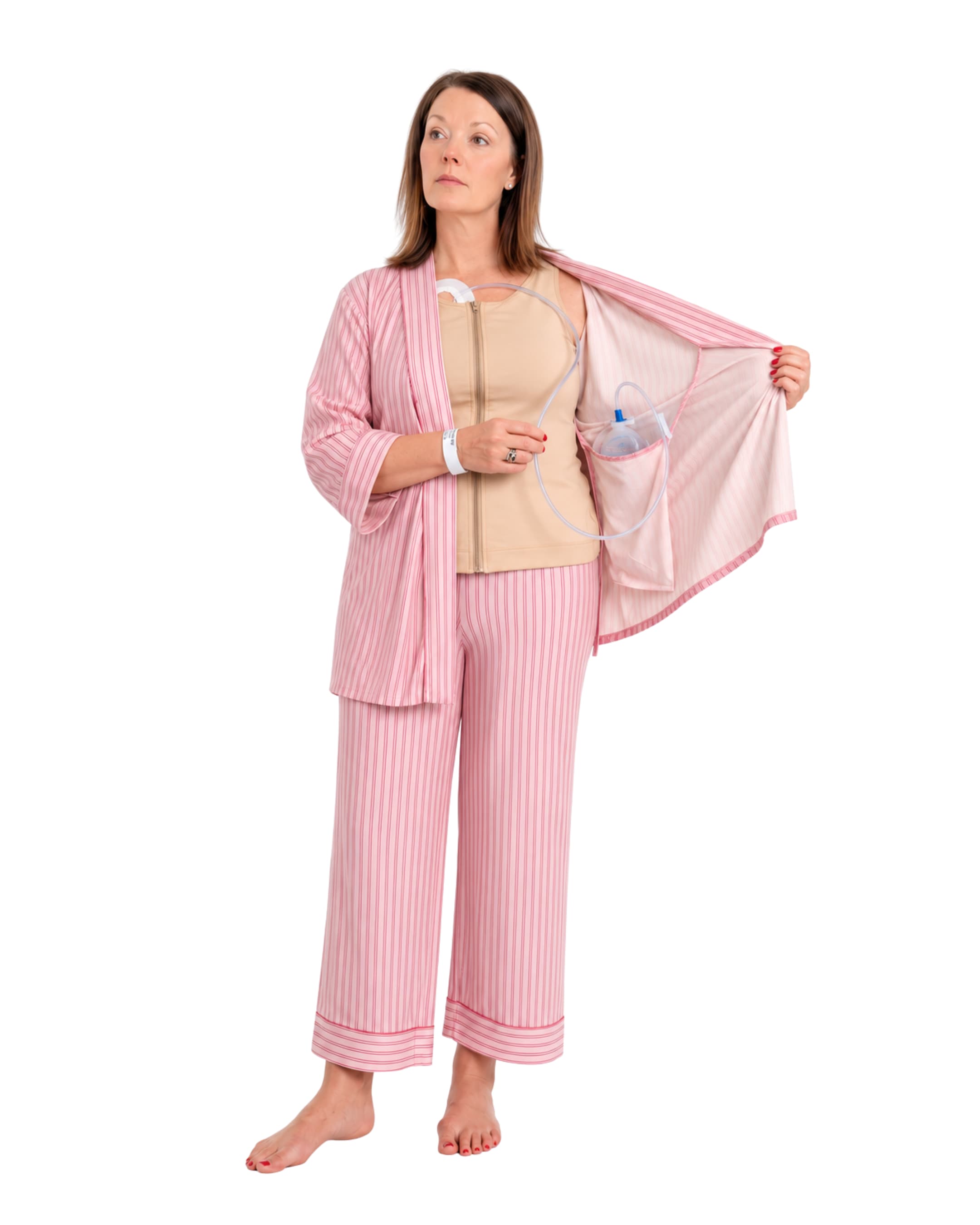A woman wearing pink striped recovery  pajamas set with and a beige mastectomy tank top, holding open the striped top while demonstrating a clear plastic  tubing and surgical jp drain placement into the interior pocket.
