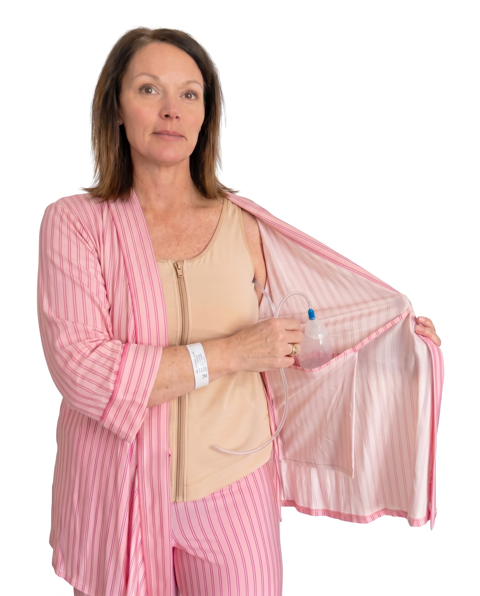 A woman wearing pink striped recovery  pajamas set with and a beige mastectomy tank top, holding open the striped top while demonstrating a clear plastic  tubing and surgical jp drain placement into the interior pocket.