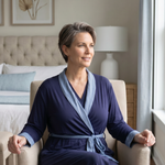 Woman sitting in a blue recovery robe with interior pockets in a blue and beige toned bedroom as she sits on a chair looking out a window post surgery