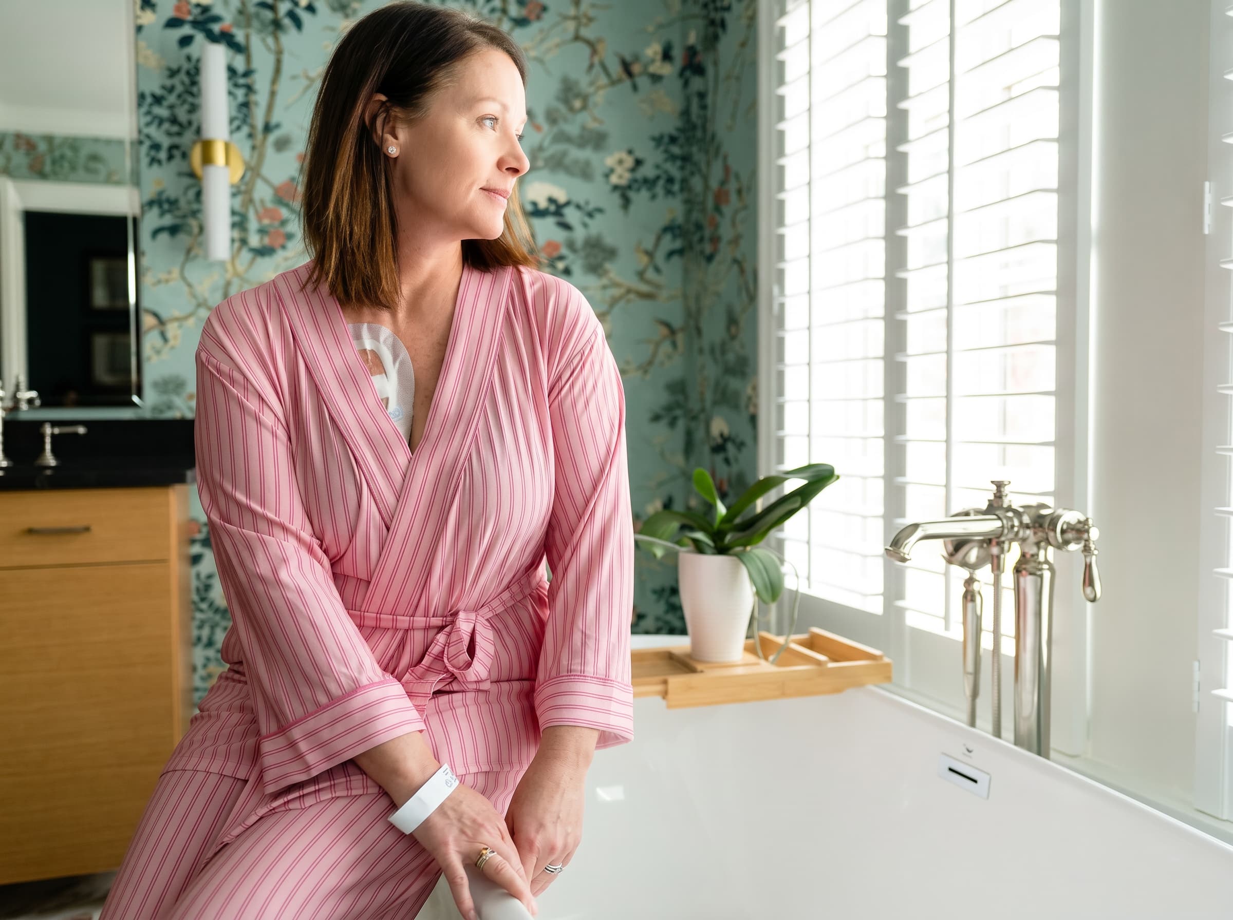 A woman wearing a vertically striped pajama robe top and matching pants sits on the edge of a white bathtub in a bathroom with floral wallpaper and white shutters.
