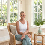 Woman sitting in a wicker chair in a bright room with large windows and plants. Woman is wearing a white zipper front opening tank top 
