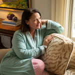 Woman in a sage green recovery robe sitting on a couch with a patterned pillow, in a cozy living room. Striped pink pajama pants peak out at the bottom