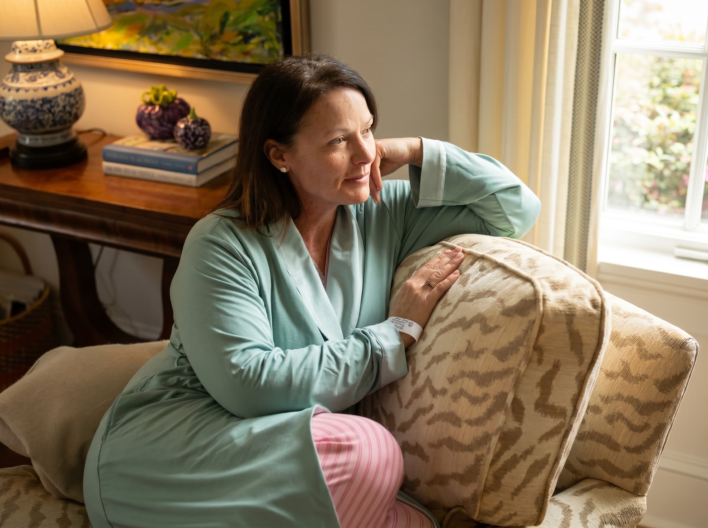 Woman in a sage green recovery robe sitting on a couch with a patterned pillow, in a cozy living room. Striped pink pajama pants peak out at the bottom