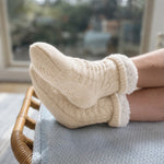 Person wearing oatmeal beige cable knitted socks with fleece lining insides and gripper bottoms sitting on a chair in a cozy living room.