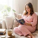 woman wearing pink striped recovery pajamas sitting on a couch reading a book