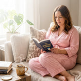 woman wearing pink striped recovery pajamas sitting on a couch reading a book