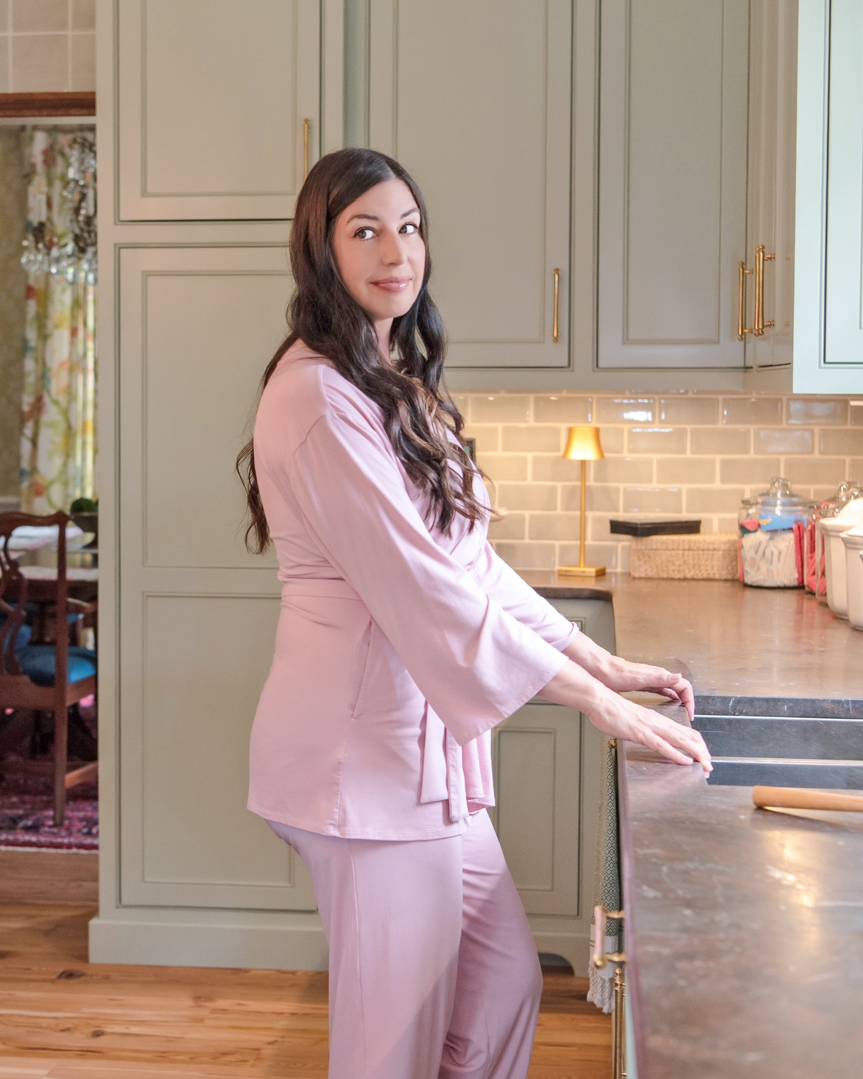 A woman with long dark hair stands in a kitchen wearing a light pink matching mastectomy shirt with drain pockets and pants set. She is leaning on a dark countertop, with light green cabinets, a subway tile backsplash, and a small table lamp in the background.