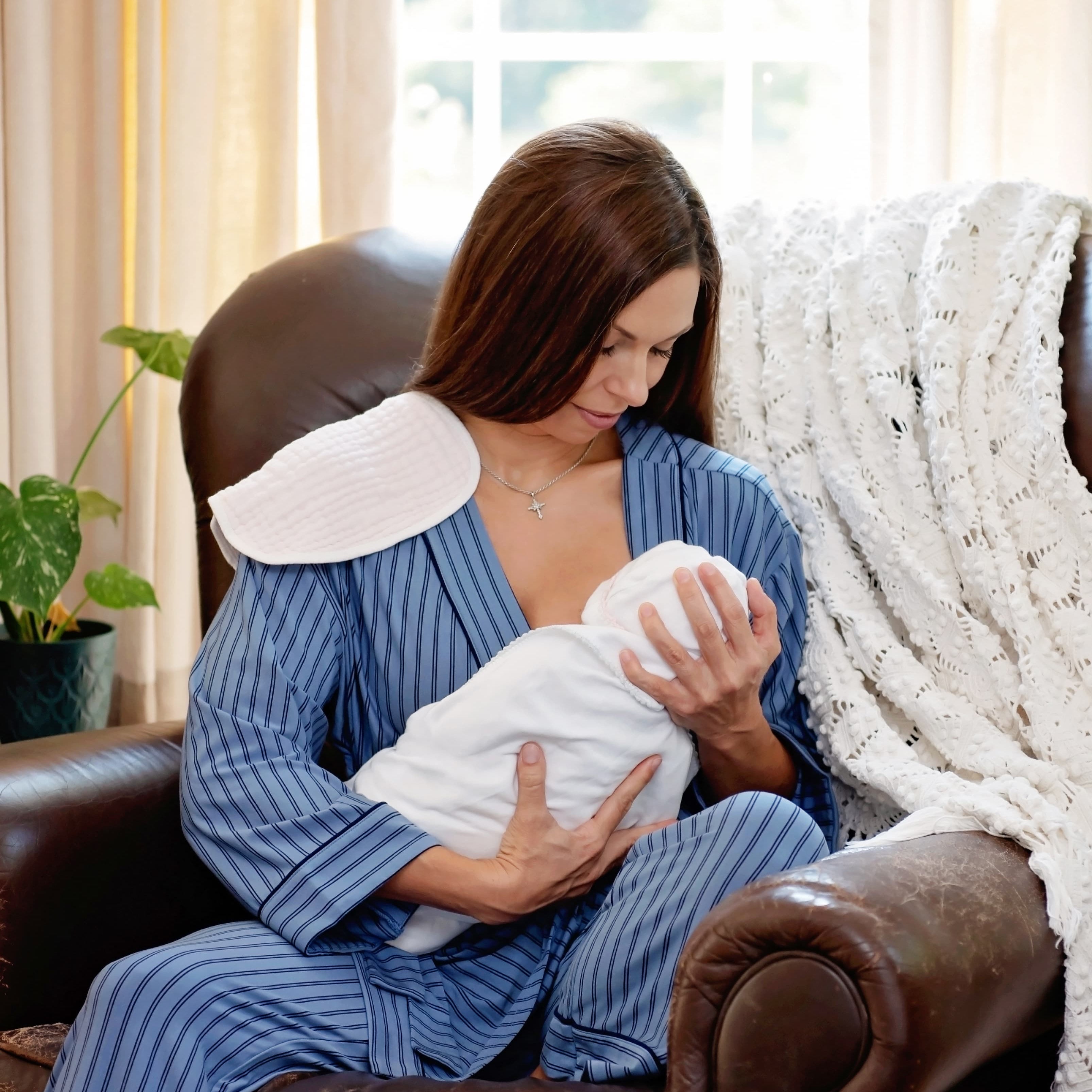 A woman wearing a blue striped wrap robe top with matching recovery pants sitting in a brown leather armchair holding a baby swaddled and nursing