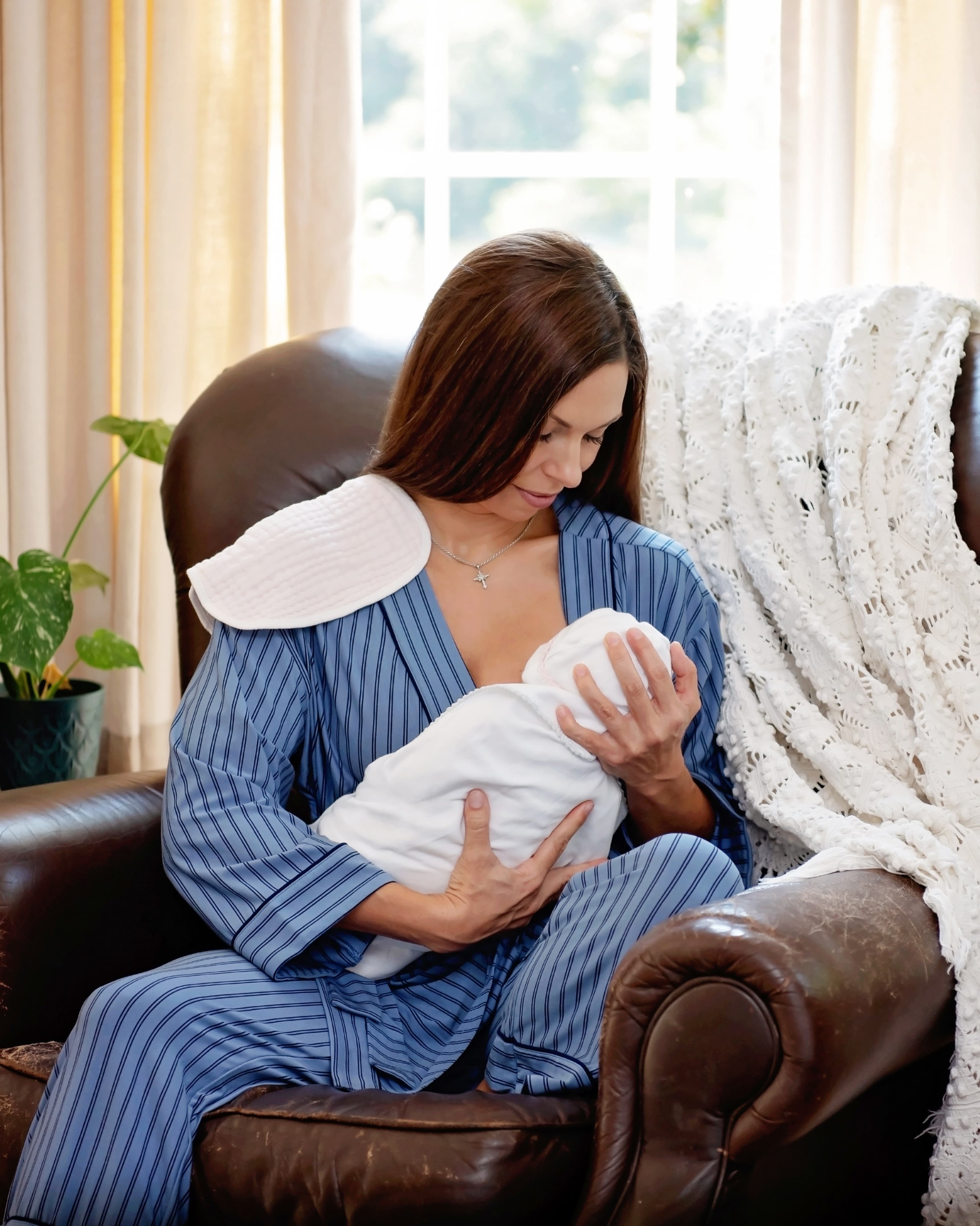 A woman wearing a blue striped wrap robe top with matching recovery pants sitting in a brown leather armchair holding a baby swaddled and nursing