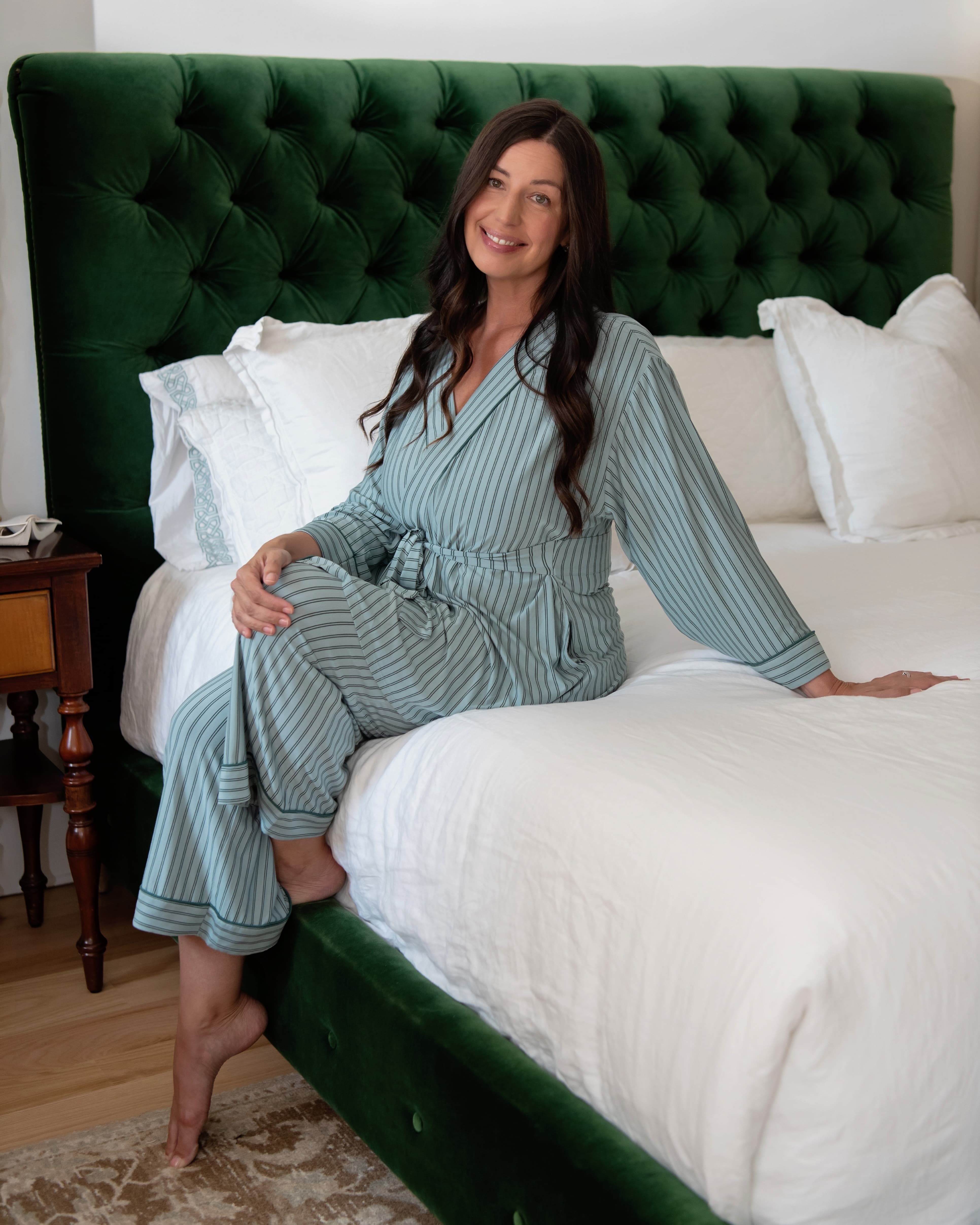 A woman in a sage green striped pajamas set sits on a bed with a green headboard and white comforter.