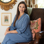 A woman with long brown hair sits in a brown leather armchair wearing a blue and gray vertically striped robe.She is recovering from surgery discreetly.