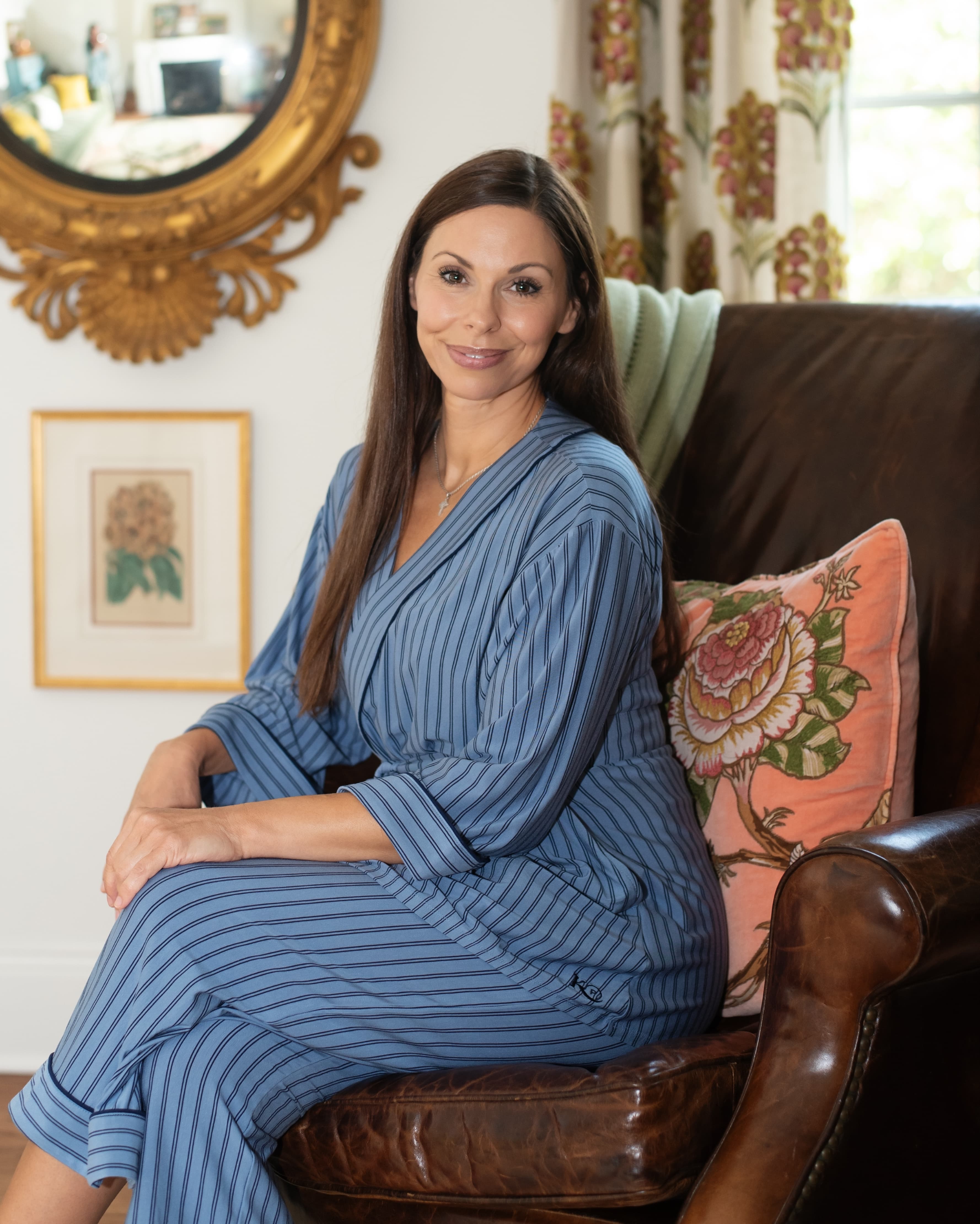 A woman with long brown hair sits in a brown leather armchair wearing a blue and gray vertically striped robe.She is recovering from surgery discreetly.
