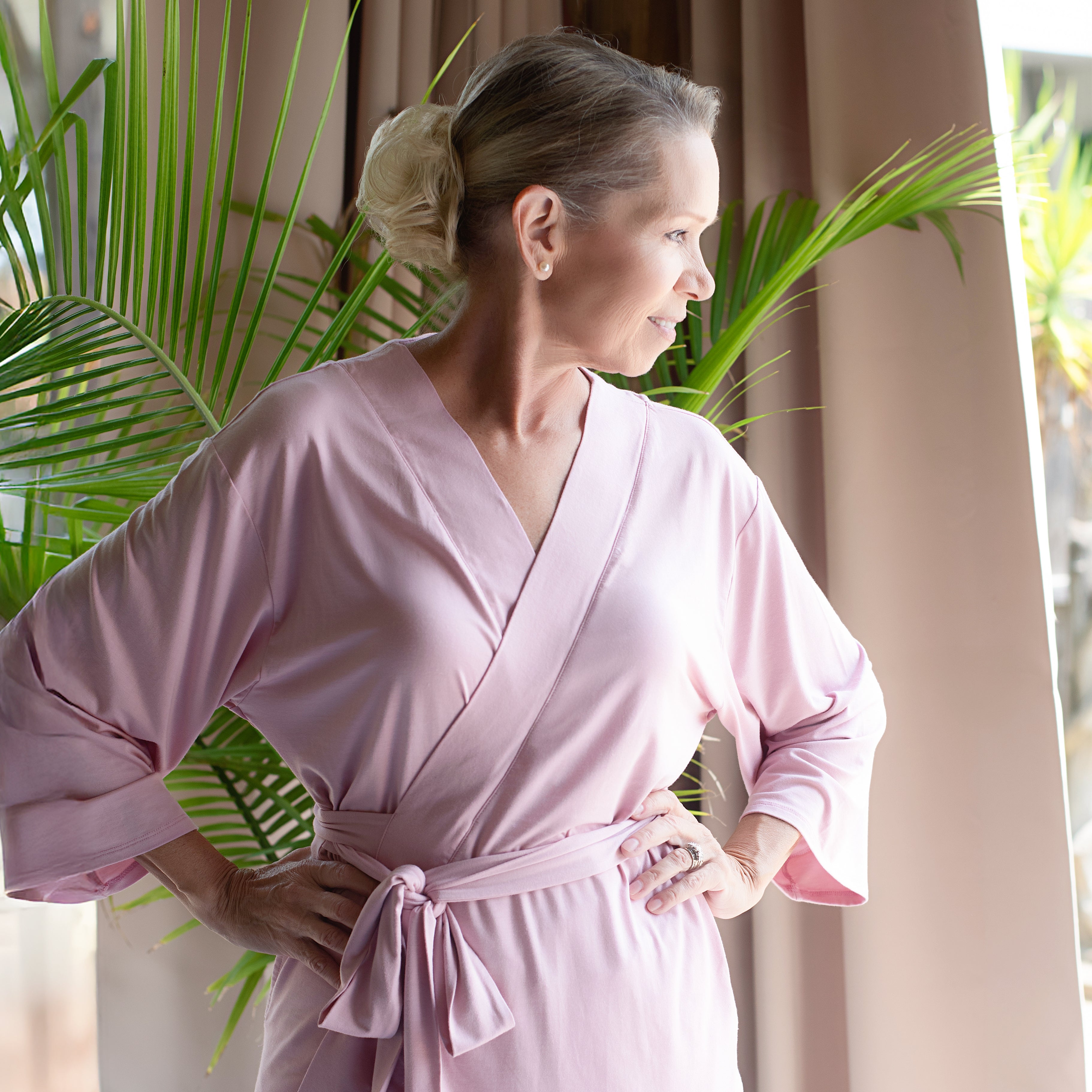 Woman wearing a pink mastectomy pajamas set standing in a room with plants and curtains.