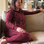 Woman sitting on a beige couch wearing a maroon recovery set in a living room with a kitchen in the background.