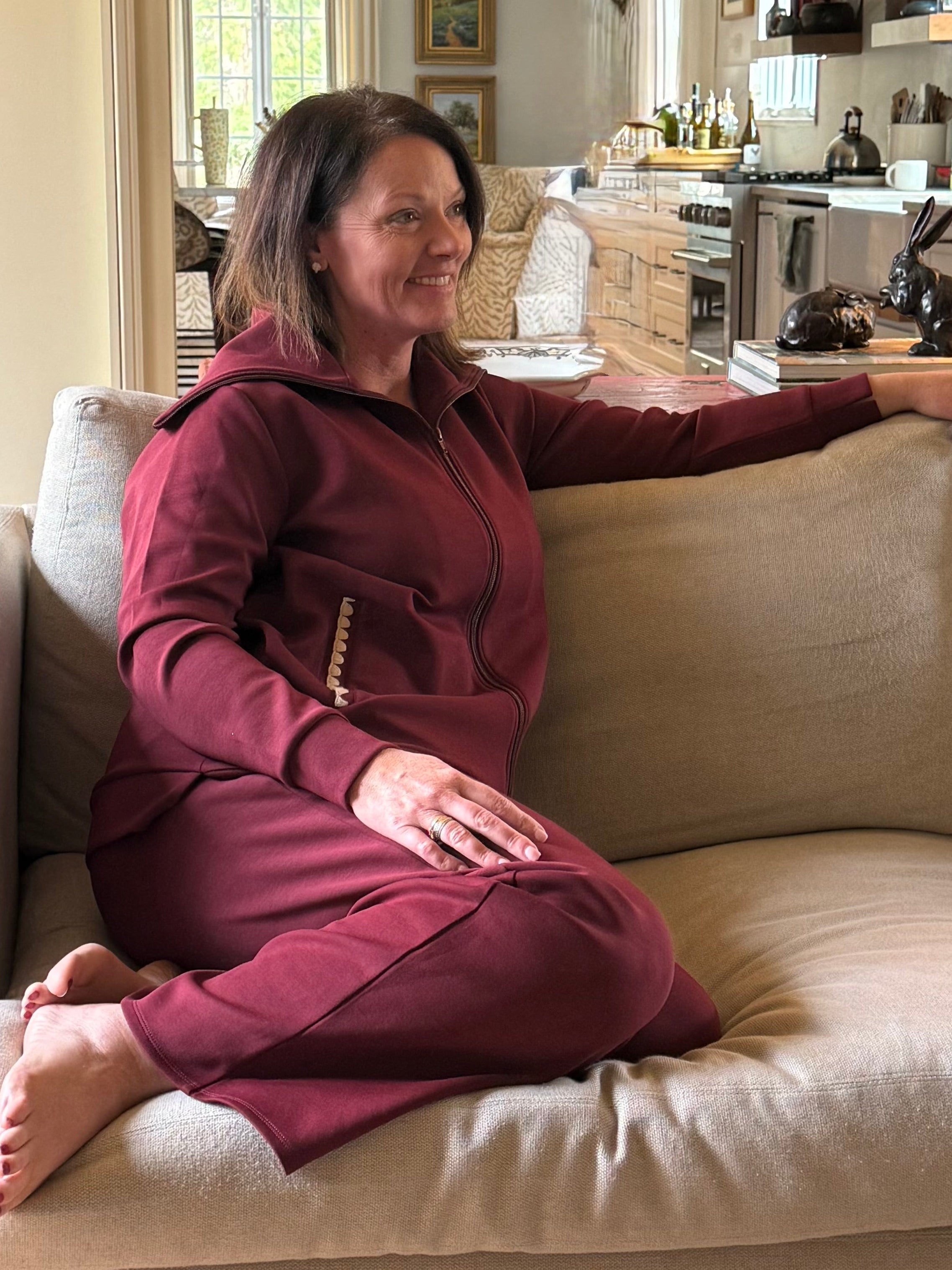 Woman sitting on a beige couch wearing a maroon recovery set in a living room with a kitchen in the background.