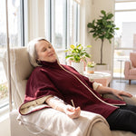 An older woman reclines in a beige treatment chair with an IV line connected to her arm, wearing a burgundy medical infusion jacket in a bright, modern treatment room with large windows and indoor plants.