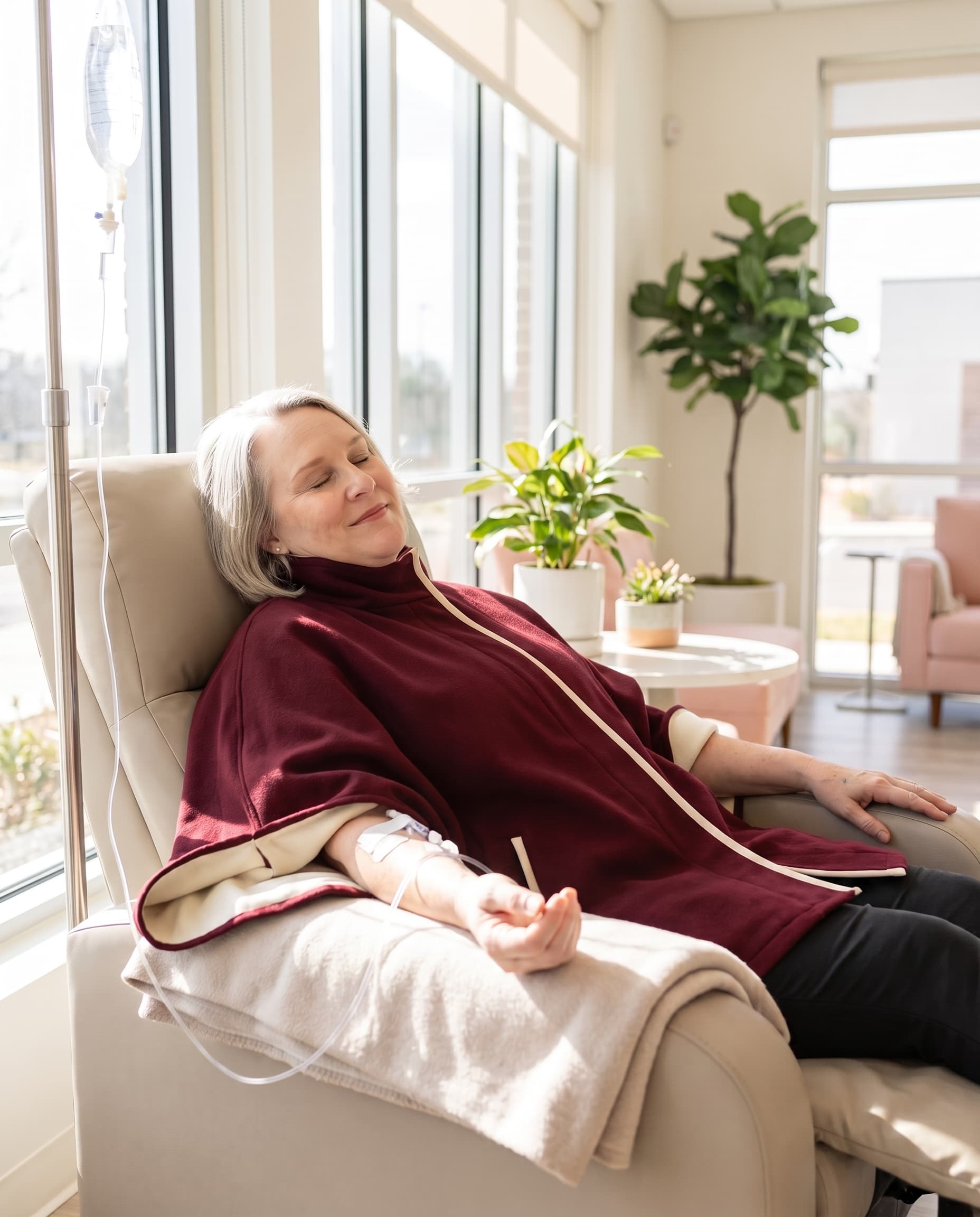 An older woman reclines in a beige treatment chair with an IV line connected to her arm, wearing a burgundy medical infusion jacket in a bright, modern treatment room with large windows and indoor plants.