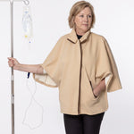A woman wearing a beige medical poncho stands next to an IV pole with an IV bag, against a plain white background.