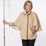 A woman wearing a beige medical poncho stands next to an IV pole with an IV bag, against a plain white background.