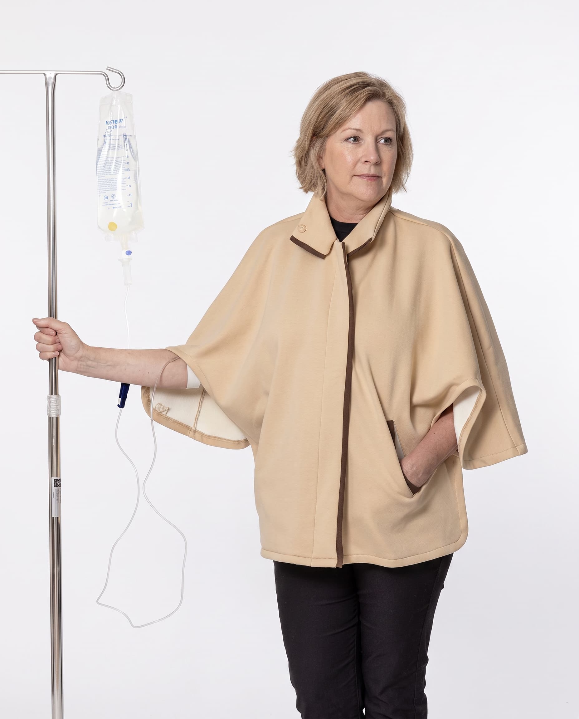 A woman wearing a beige medical poncho stands next to an IV pole with an IV bag, against a plain white background.
