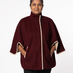 A woman wearing a burgundy cape-style jacket with cream-colored trim on the front placket, sleeve cuffs, and side pockets, photographed against a plain white studio background.