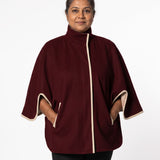 A woman wearing a burgundy cape-style jacket with cream-colored trim on the front placket, sleeve cuffs, and side pockets, photographed against a plain white studio background.
