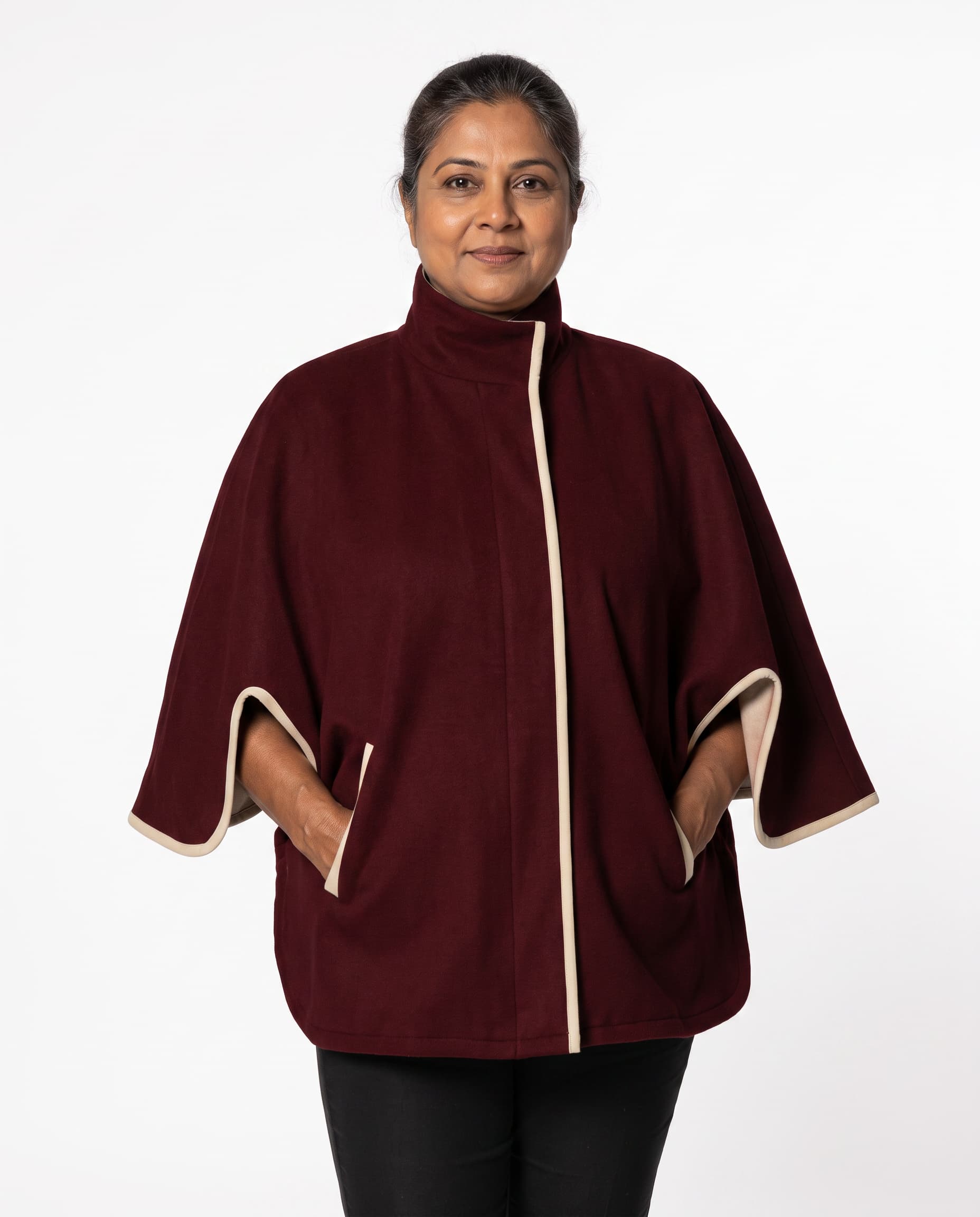 A woman wearing a burgundy cape-style jacket with cream-colored trim on the front placket, sleeve cuffs, and side pockets, photographed against a plain white studio background.