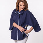 A woman wearing a navy blue poncho-style top with white piping trim buttoning the underarm button that highlights the adaptive function of the cape so that it can come off and be put on without detaching from medical equipment, shown against a plain light gray studio background.
