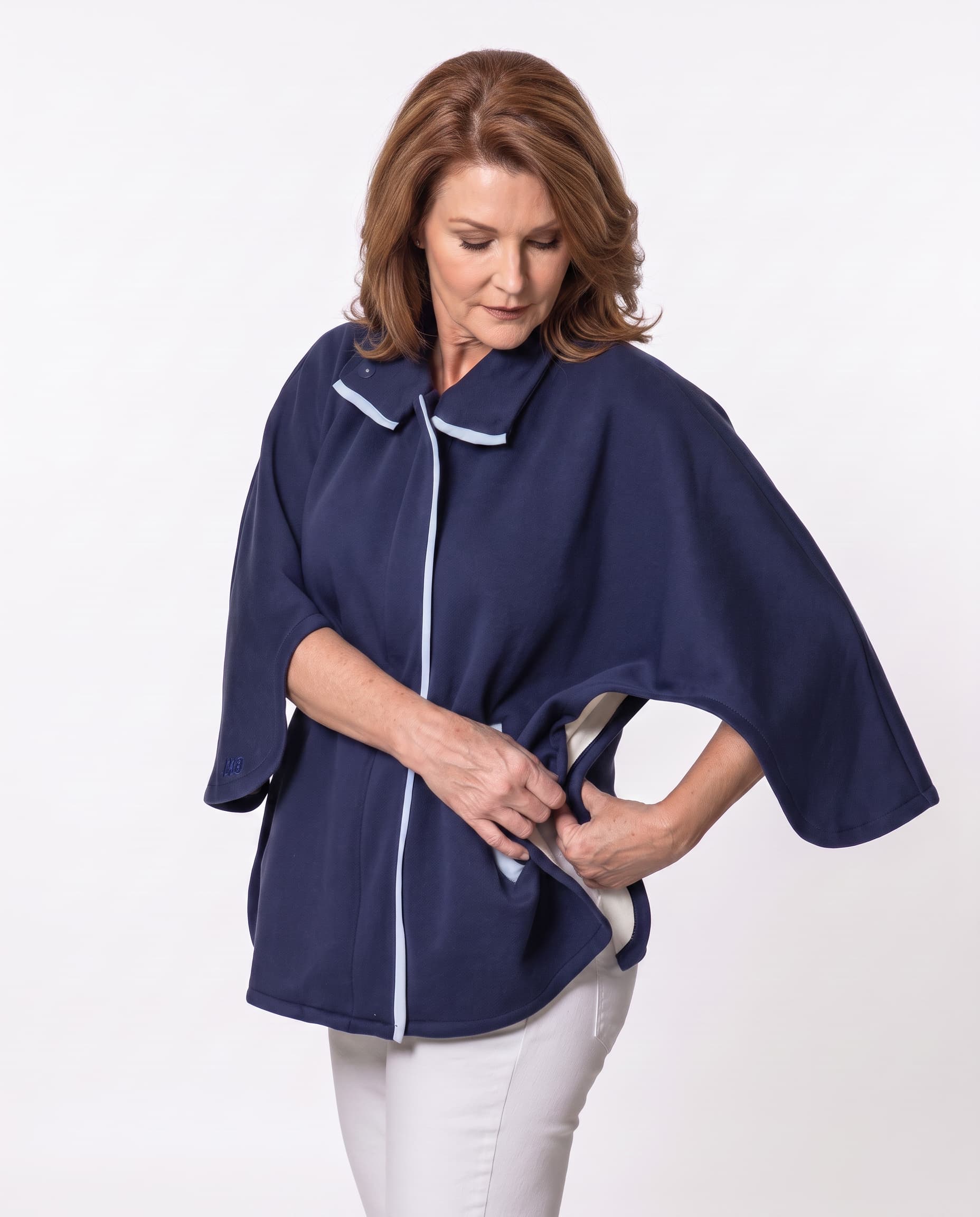 A woman wearing a navy blue poncho-style top with white piping trim buttoning the underarm button that highlights the adaptive function of the cape so that it can come off and be put on without detaching from medical equipment, shown against a plain light gray studio background.