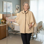 A smiling woman stands in a treatment room wearing a beige cape-style top over black pants, with an IV pole at her side and hospital beds in the background.