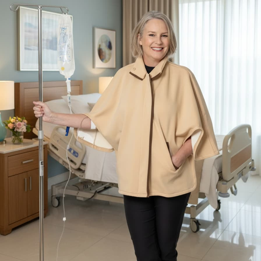 A smiling woman stands in a treatment room wearing a beige cape-style top over black pants, with an IV pole at her side and hospital beds in the background.