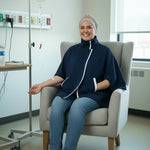 A smiling woman wearing a navy blue medical poncho with white trim, blue jeans, and white sneakers sits in a light gray armchair inside a hospital room. An IV pole and medical wall outlets are visible in the background.