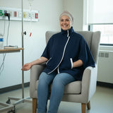 A smiling woman wearing a navy blue medical poncho with white trim, blue jeans, and white sneakers sits in a light gray armchair inside a hospital room. An IV pole and medical wall outlets are visible in the background.