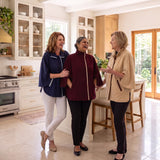 Three women standing and conversing in a bright, modern kitchen all wearing the KickIt Infusion Jacket, one in beige, one in maroon and one in navy. The maroon jacket is zipped up to highlight the turtle neck while the camel and navy are slightly unzipped featuring the collar look.