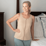 Woman wearing a camel sleeveless mastectomy tank top that zippers up the front, dual-directions and light green pants standing in a bedroom.