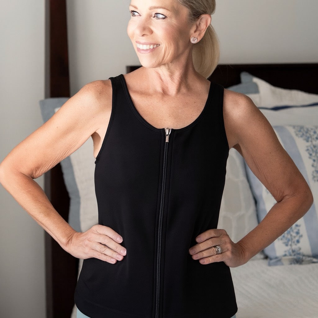 woman wearing the mastectomy tank top in black with it zipped up at the front standing next to a bed