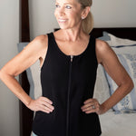 woman wearing the mastectomy tank top in black with it zipped up at the front standing next to a bed