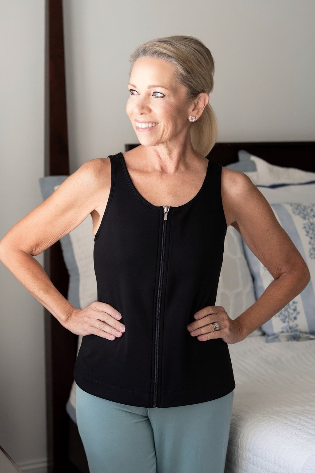 woman wearing the mastectomy tank top in black with it zipped up at the front standing next to a bed
