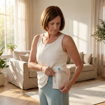 Woman wearing a white mastectomy tank top holding out the interior drain pocket to highlight the surgical drain bulb against a living room background