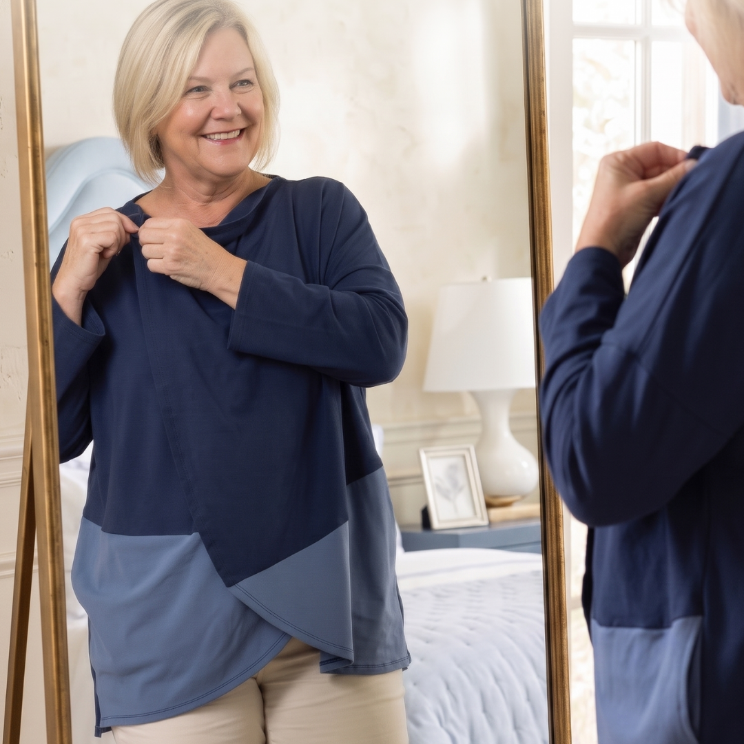 A woman wearing a navy and blue color-block long-sleeve wrap top, seen both in front of and reflected in a gold-framed mirror in a bedroom setting to highlight the snap closure at the neck for someone with limited mobility