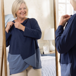 A woman wearing a navy and blue color-block long-sleeve wrap top, seen both in front of and reflected in a gold-framed mirror in a bedroom setting to highlight the snap closure at the neck for someone with limited mobility