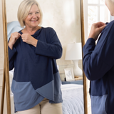 A woman wearing a navy and blue color-block long-sleeve wrap top, seen both in front of and reflected in a gold-framed mirror in a bedroom setting to highlight the snap closure at the neck for someone with limited mobility