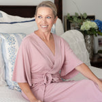 Woman in a pink robe and adaptive pants pajamas set sitting on a bed with a floral arrangement in the background